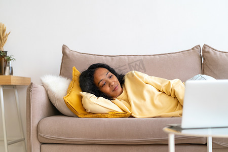 美国客厅摄影照片_年轻的非洲裔美国女性躺在客厅的沙发上，在笔记本电脑上看电影