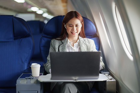 亚洲女商人摄影照片_一位成功的亚洲女商人或女性企业家在飞机上穿着正式西装的肖像坐在商务舱座位上，并在飞行期间使用笔记本电脑