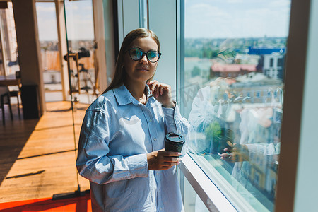 一位戴眼镜的年轻女性自由职业者端着咖啡站在一扇大窗户旁，看着这座城市。