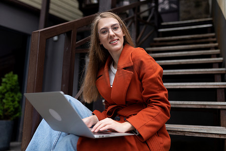 坐在城市楼梯上、穿着秋装的年轻女性在笔记本电脑上在线工作