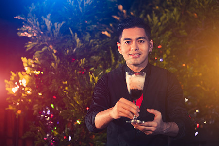 年轻的亚洲男性调酒师在户外夜总会将酒精饮料鸡尾酒与玻璃酒混合。