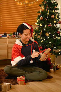 戴着圣诞老人帽子的男人打开圣诞礼物，坐在装饰好的房间里。