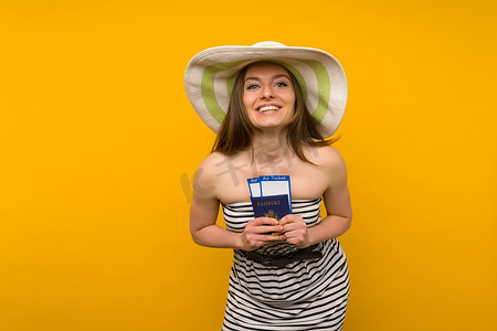 戴着草帽和条纹连衣裙的快乐的年轻女子拿着黄色背景护照的机票。