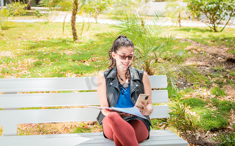 一个女孩坐着用笔记本和手机做笔记，一个女人坐着用手机做笔记，拉丁女孩在公园里用手机