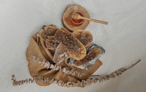 美味的蜂蜜从玻璃罐上的新鲜蜂窝中滴落下来，木制蜂蜜勺子放在麻布上。