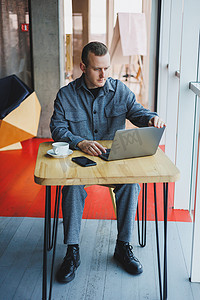 一位身穿西装的年轻成功商人喝着咖啡，在咖啡馆里用笔记本电脑无线免费上网工作，独自坐在咖啡馆的桌子旁。