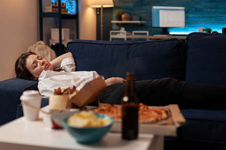 手绘披萨插画摄影照片_在电视前喝啤酒和吃外卖披萨后睡在沙发上的人