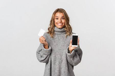 身穿灰色毛衣的漂亮金发女孩，在白色背景下展示带信用卡的智能手机屏幕，用于网上购物