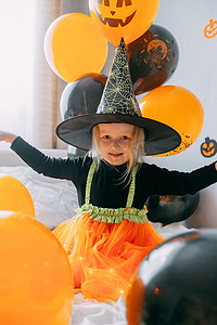 一个女巫摄影照片_儿童万圣节-一个戴着女巫帽的女孩和一个狂欢节服装，家里有通风的橙色和黑色气球。