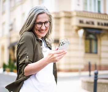 成熟美丽的灰发女人看着智能手机走在古老的欧洲小镇的街道上。