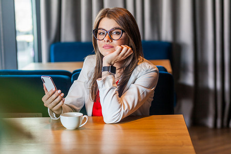 咖啡厅女孩摄影照片_女人手里拿着一杯咖啡和智能手机，表情平静地看着相机。