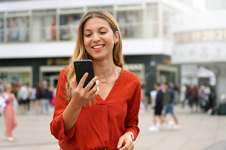 应用视频摄影照片_德国柏林城市广场，手持手机观看应用视频的迷人年轻女性