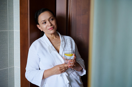 清爽浴室摄影照片_穿着白色华夫格浴袍的美丽中年多民族妇女，配上一杯新鲜的柠檬水。