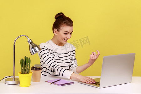 女经理坐在工作场所，向笔记本电脑屏幕展示并做出招手的手势。