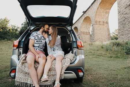 中年夫妇坐在 SUV 汽车后备箱内，背景是高架桥，白人男女一起享受公路旅行