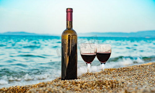 红色海浪摄影照片_绿松石海浪背景下海滩上的一瓶酒和玻璃杯