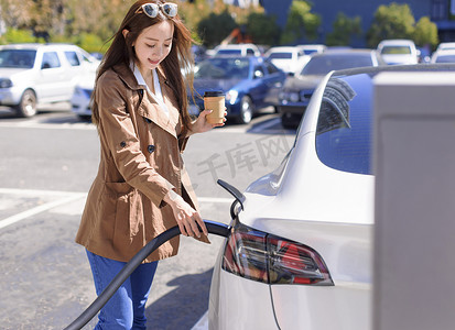 微笑的年轻女子站在电动车附近的城市停车场，从小城市车站为汽车电池充电