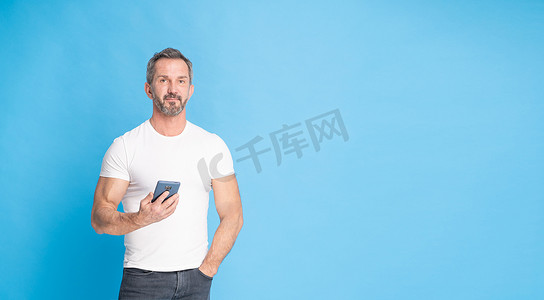 40 多岁的英俊中年灰发男子站着，手里拿着智能手机，身穿蓝色背景的白色 T 恤。