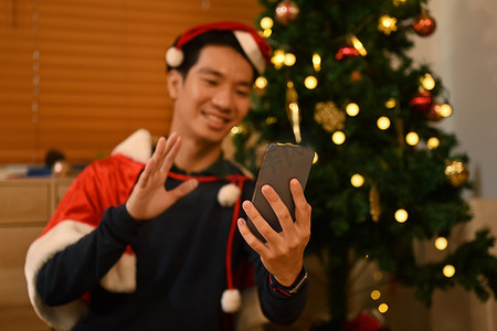戴着圣诞帽的快乐男人坐在客厅里用智能手机进行视频通话，客厅里装饰着灯光花环