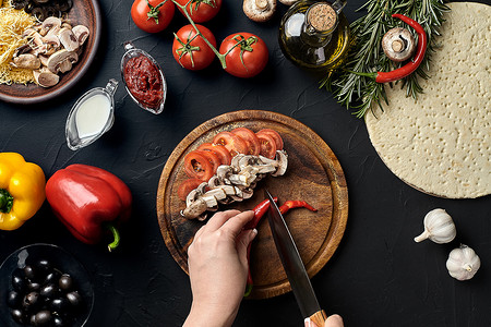 厨房桌子上的木板上放着女性手切的辣椒、蘑菇和西红柿，周围是披萨的配料：蔬菜、奶酪和香料。
