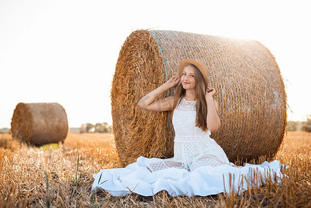 夕阳下的人摄影照片_穿着白色裙子的快乐女孩和一顶草帽坐在收获麦田的稻草包下