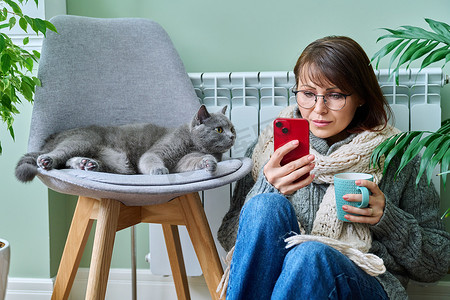 在家使用智能手机的女性，在暖气片附近用猫取暖