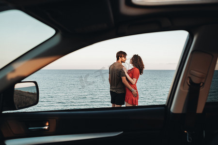 透过车窗看年轻夫妇站在海的背景上，日落时男人拥抱穿着红裙的卷发女人