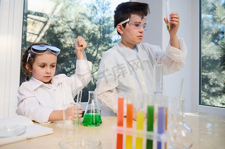 化学实验室穿着摄影照片_穿着白色实验室外套和防护眼镜的可爱男孩和​​女孩，在化学课上进行生物化学研究