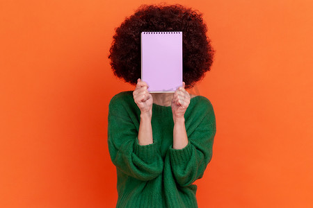 藏风格摄影照片_身穿绿色休闲风格毛衣的黑人发型不知名女性的画像站着拿着组织者，把脸藏在纸质笔记本后面。