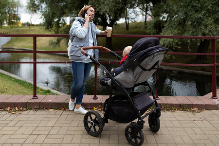 年轻有爱心的母亲在公园里散步时，手里拿着一杯咖啡，坐在婴儿车旁边，带着刚出生的孩子，脸上挂着微笑，在电话里说话