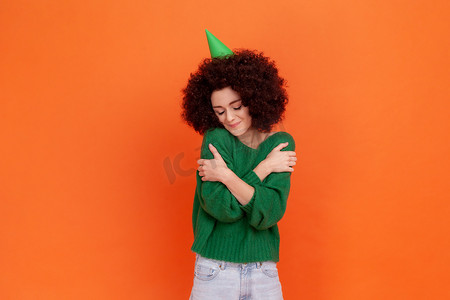 善良的生日女人，留着非洲发型，穿着绿色休闲风格的毛衣和派对锥，庆祝节日，拥抱自己，利己主义者。