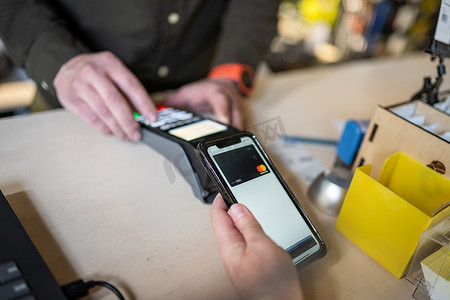 裁剪视图的女人站在收银台，支付在商店购物，使用现代智能手机，而收银员手持 NFC 终端。