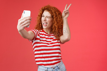 时髦时髦俏皮好看的红发大胆卷发女人展示和平胜利动物耳朵手势眨眼智能手机显示记录视频博客自拍真棒手机相机红色背景