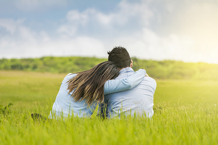 坐在草地摄影照片_浪漫的情侣坐在草地上从后面拥抱，一对相爱的情侣在草地上拥抱的后视图，一对相爱的情侣坐在草地上从后面拥抱