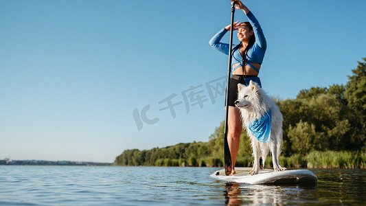 清晨，快乐的年轻女子和她的狗雪白的日本狐狸站在 Sup 板上享受湖边的生活