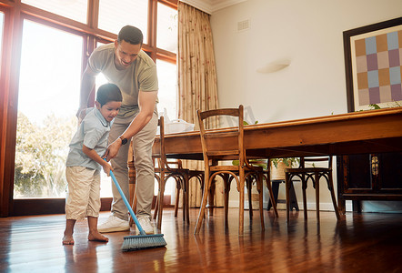 男孩家务摄影照片_混血父亲帮助小男孩用扫帚扫木地板上的灰尘和污垢，做家务。