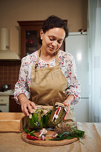 宜人的女人，一位身穿米色厨师围裙的家庭主妇，在自家厨房里摆着自制的腌制时令蔬菜