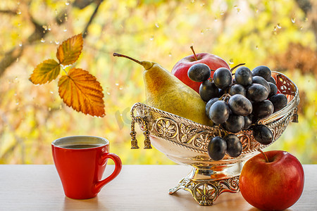 水滴梨摄影照片_带梨、葡萄和苹果的金属花瓶，窗玻璃上的秋黄叶，模糊的背景中有水滴