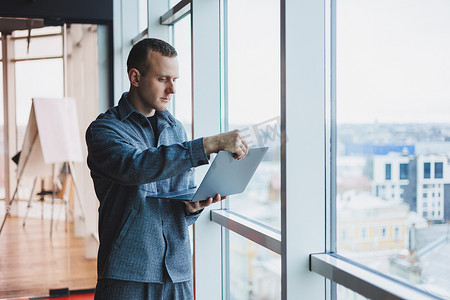 一个聪明聪明的男性经理的肖像，他拿着一台笔记本电脑在办公室进行研究，一个穿着优雅衣服的商人在触摸板上玩。