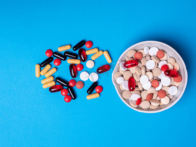 蓝色背景上一碗药丸的顶部视图，旁边是不同颜色和形状的其他药丸和胶囊