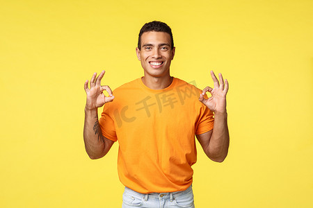 迷人的快乐男人，手臂上有纹身，身穿橙色 T 恤、白色裤子、表现不错、确认或良好手势、微笑点头同意、推荐产品、在黄色背景下推广公司