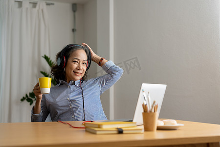 戴着耳机的快乐亚洲老年女性在舒适的家客厅里用笔记本电脑听歌，看着相机。