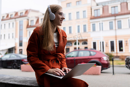 城里一名身穿秋衣的年轻女学生在笔记本电脑上在线工作，并通过耳机听音乐