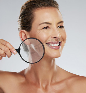 护肤、美容和成熟女性用放大镜在灰色样机工作室背景下检查脸部。