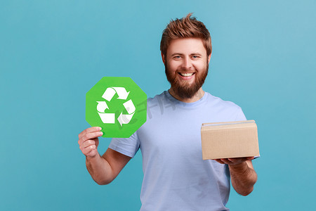 卡通生态摄影照片_身穿 T 恤的男子拿着绿色回收标志和卡通包裹，带着露齿微笑看着相机