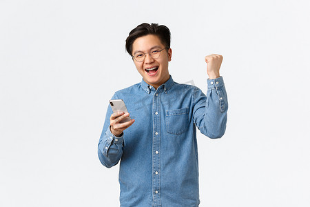 成功获胜的亚洲男子戴着眼镜欢欣鼓舞，拳头泵得满意，微笑着达到终点，玩手机游戏或在约会应用程序上约会，在白色背景下胜利喜悦