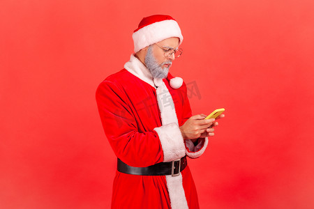 穿着圣诞老人服装、留着灰色胡须的严肃老人使用面部表情集中的智能手机、浏览互联网、社交网络。