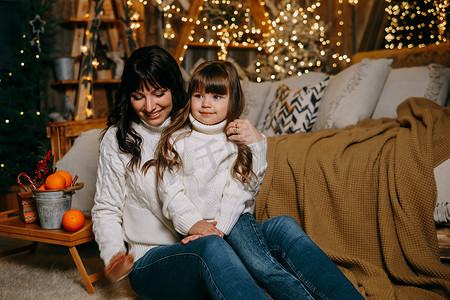 家庭环境摄影照片_一个小女孩和她的母亲在舒适的家庭环境中，坐在圣诞树旁边的沙发上。