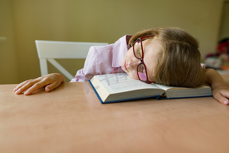 戴眼镜的小学生睡在一张桌子上，她的头放在一本打开的书上。