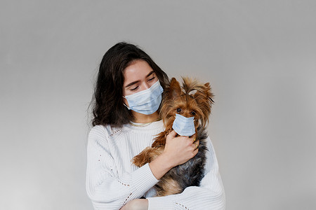 戴着外科口罩的年轻女子也抱着她戴着防护面具的狗约克夏犬。